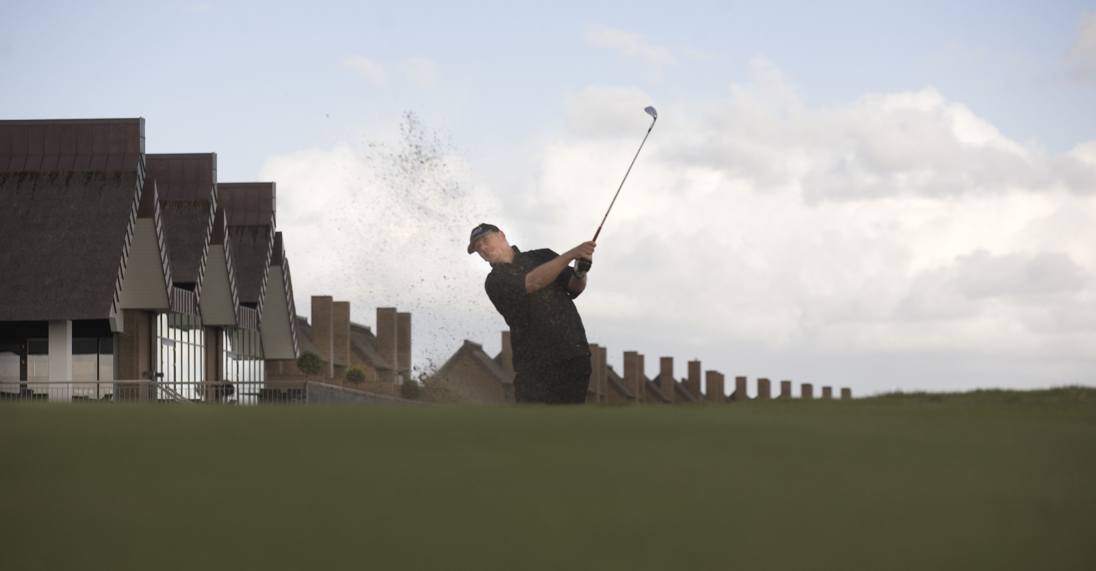 A golfer in mid-swing on a golf course with modern buildings in the background.