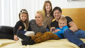A family of five on a bed with a wooden headboard, white curtains, and a painting on the wall.