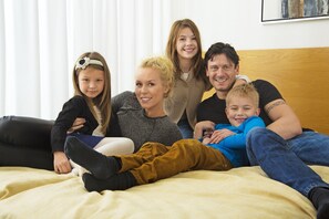 A family of five on a bed with a wooden headboard, white curtains, and a painting on the wall.