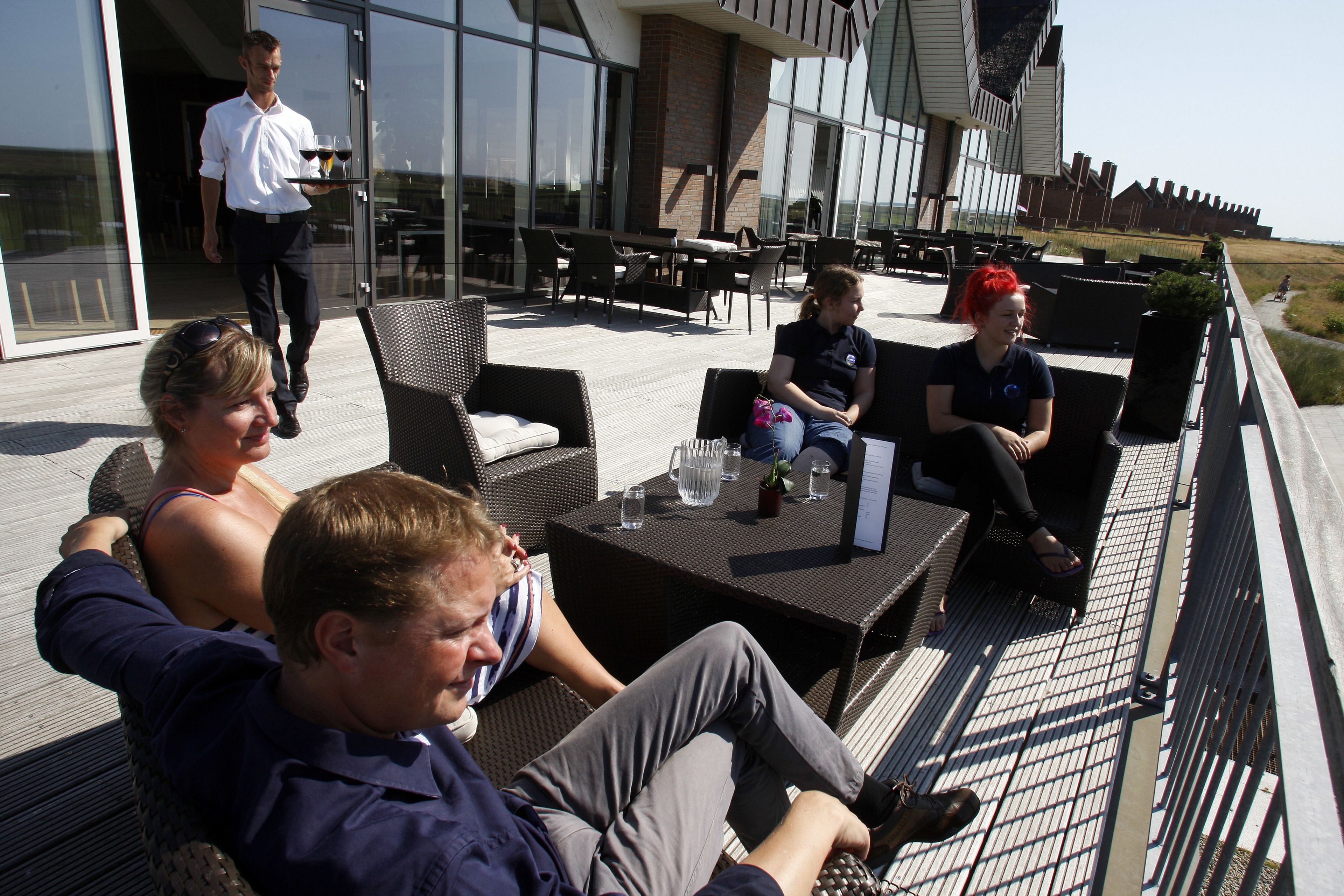 Een terras buiten met rieten meubilair en een glazen balustrade met uitzicht op een strand.
