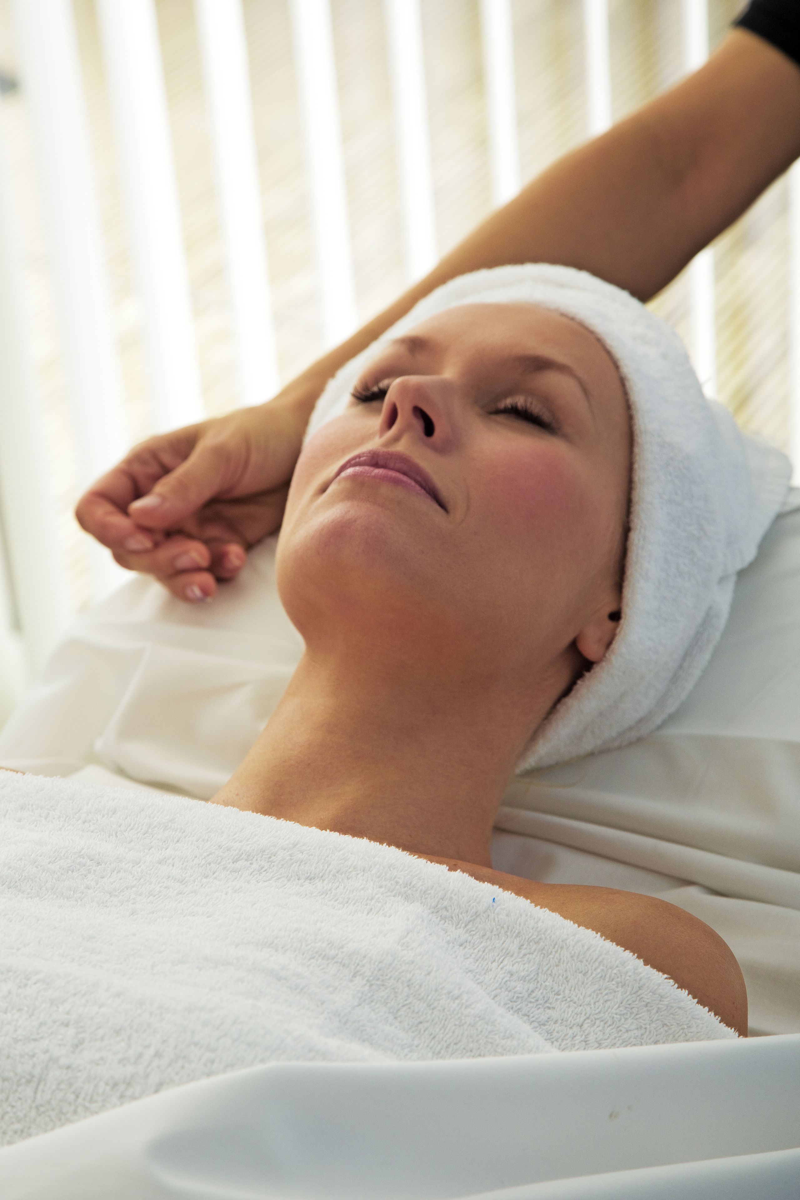A person lying on a bed with a white towel wrapped around their head, in a room with white bedding and vertical blinds in the background.
