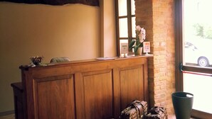 A reception desk with a wooden finish, a window with a view of the outside, and a tiled floor. - Dimora Cortese (Castagnole delle Lanze)