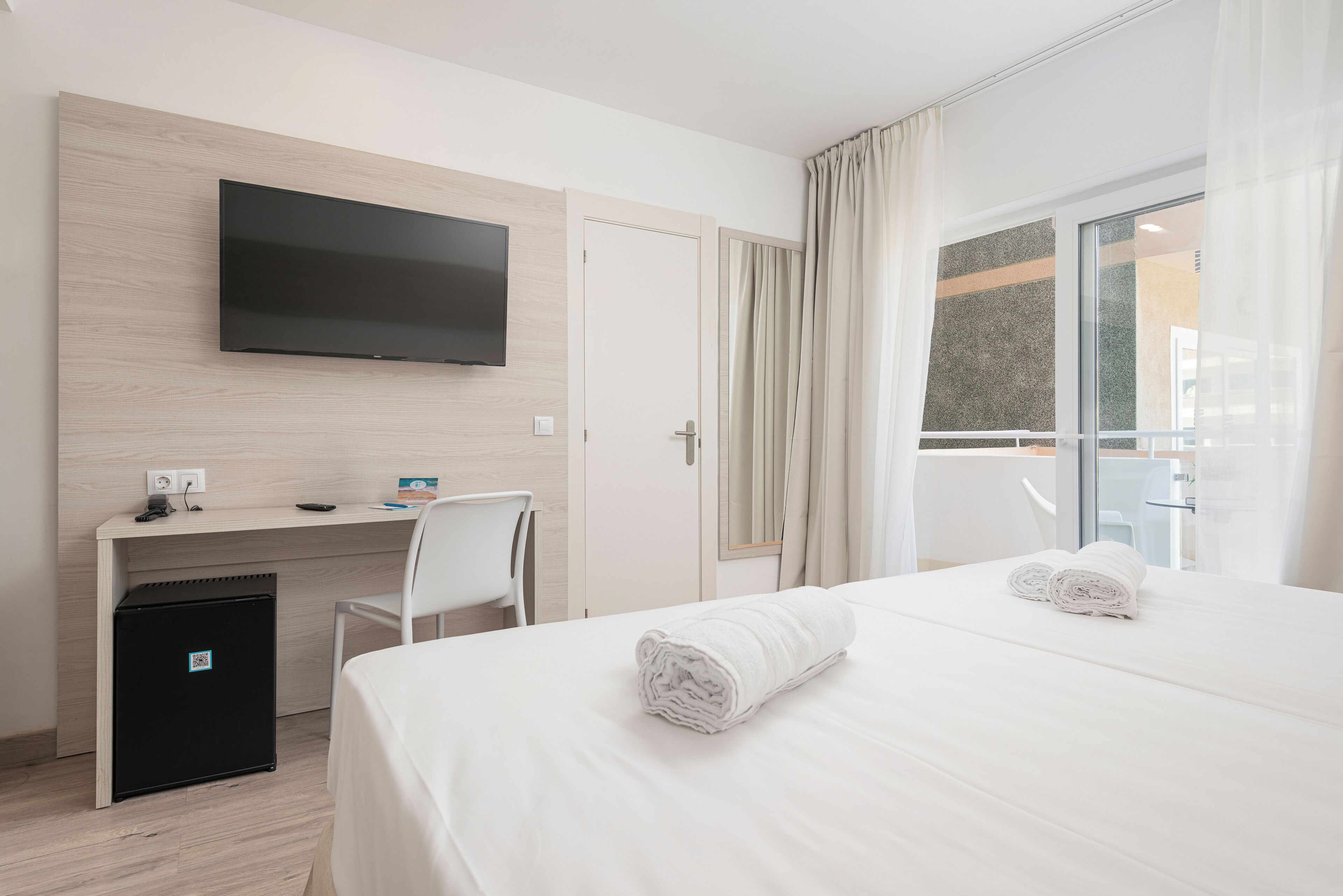 Habitación de hotel con cama, escritorio, televisión, refrigerador pequeño y ventana con cortinas.