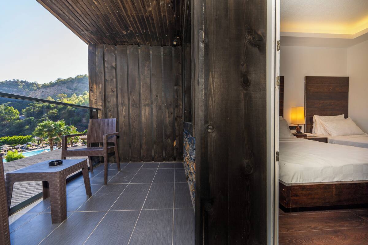 A balcony with a view of a pool and hills, a wooden wall, a bed with a headboard, and a nightstand with a lamp.