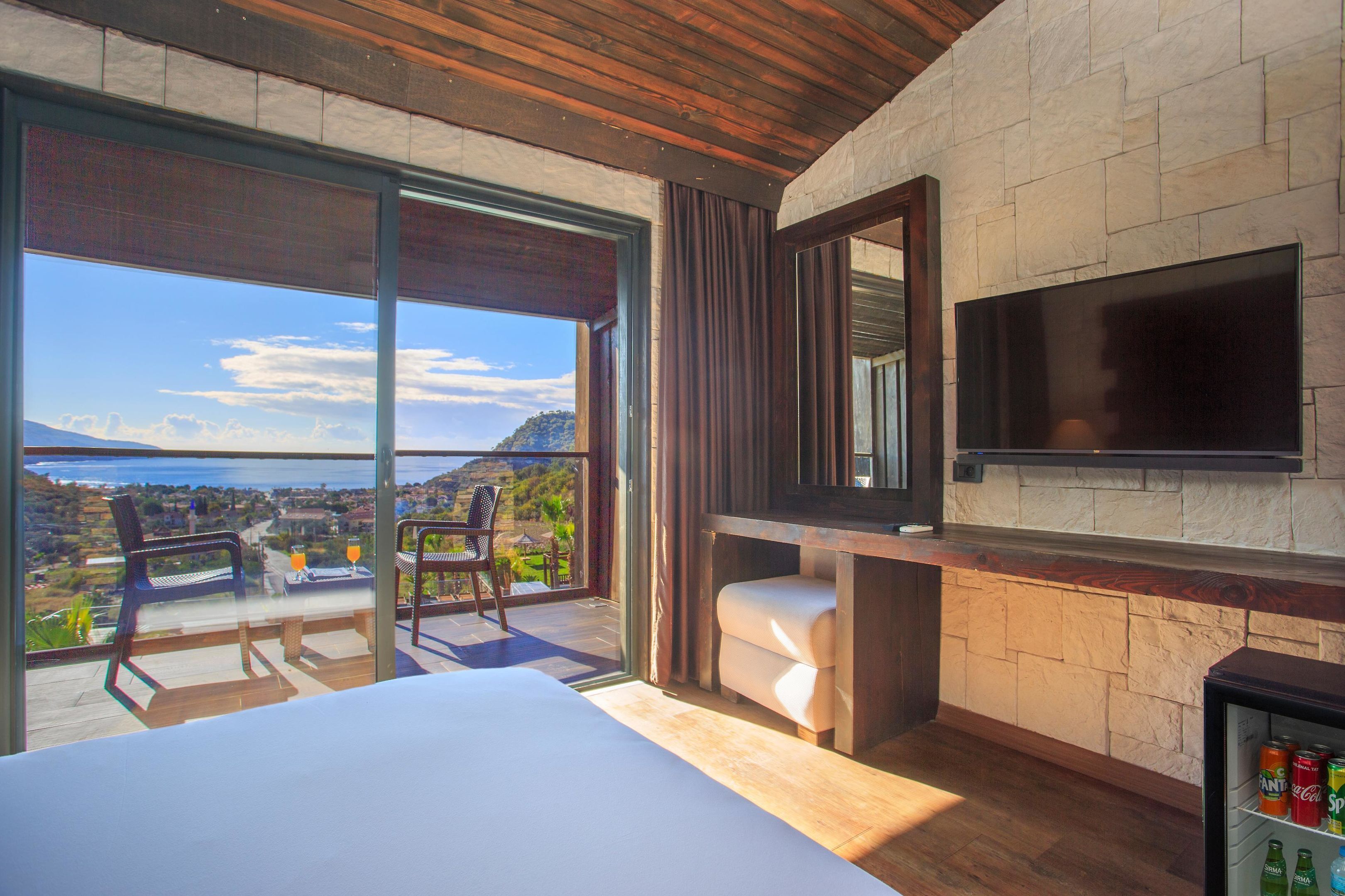 A bedroom with a large bed, a TV mounted on a stone wall, and a balcony with a view of the sea.