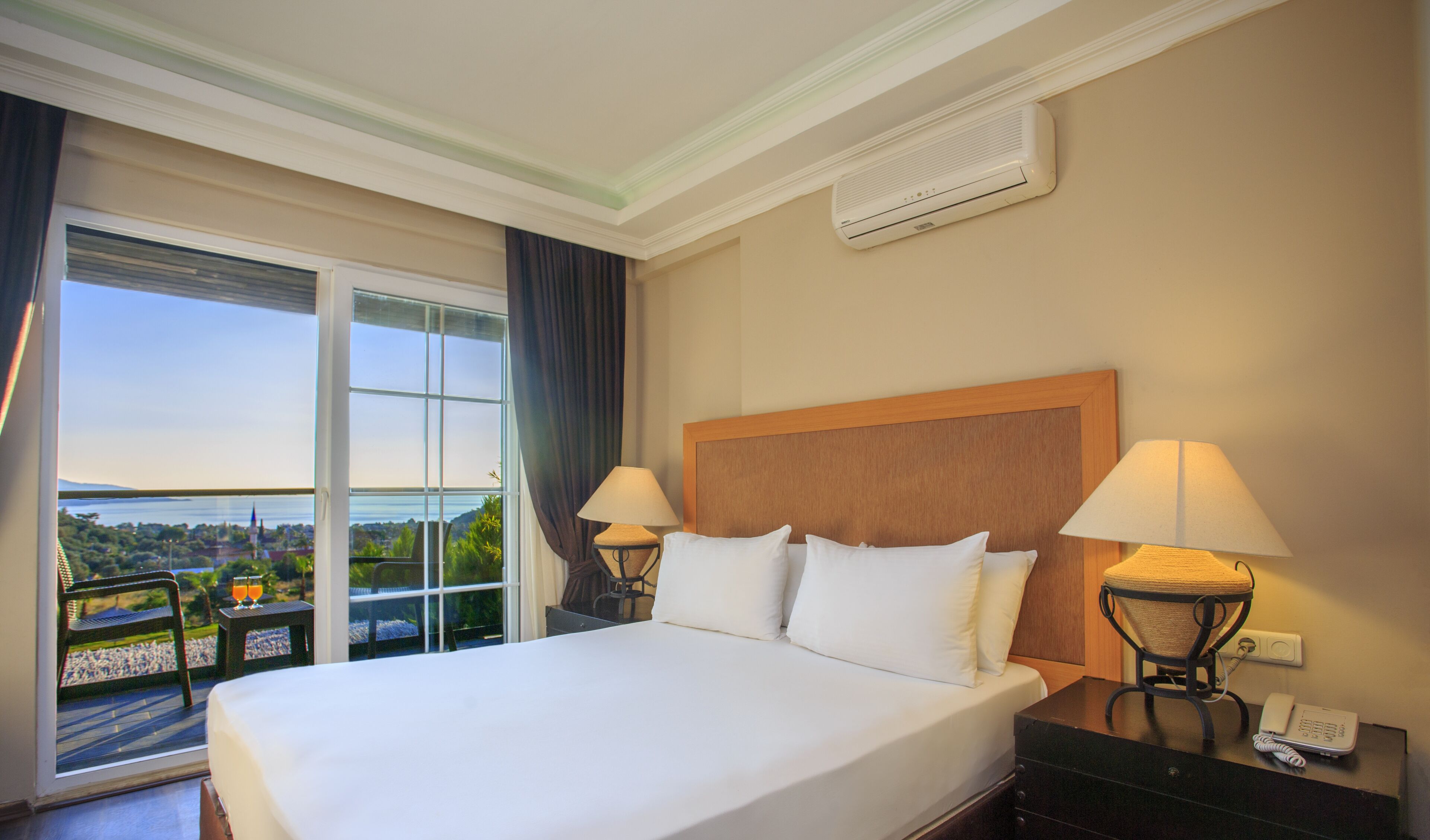 A hotel room with a large bed, two bedside lamps, a phone, and a view of a cityscape through a large window.