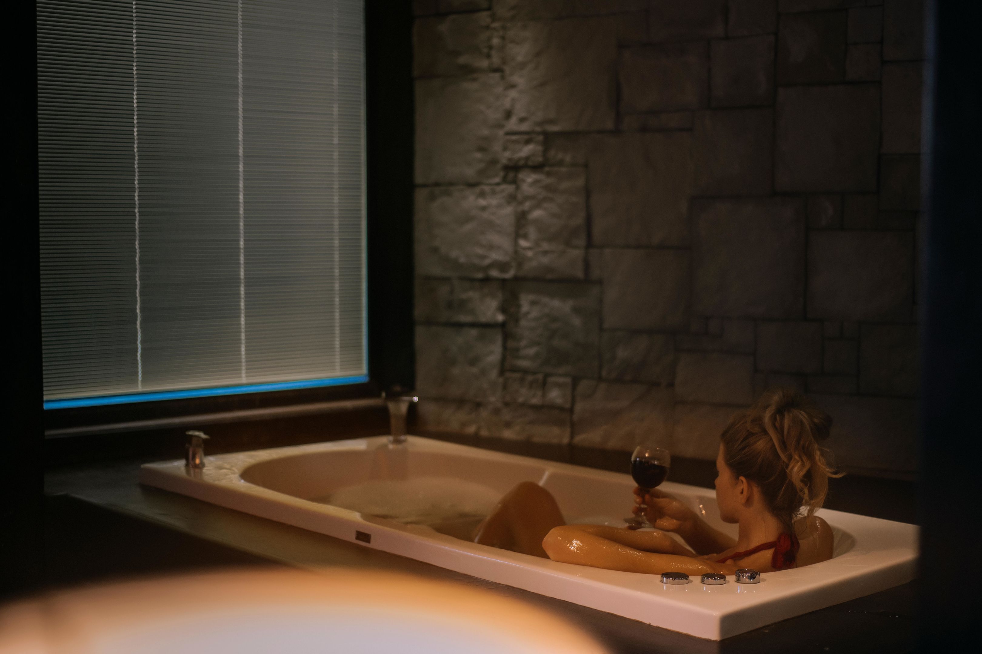 A woman in a bathtub with a wine glass, a window with blinds, and a stone wall.