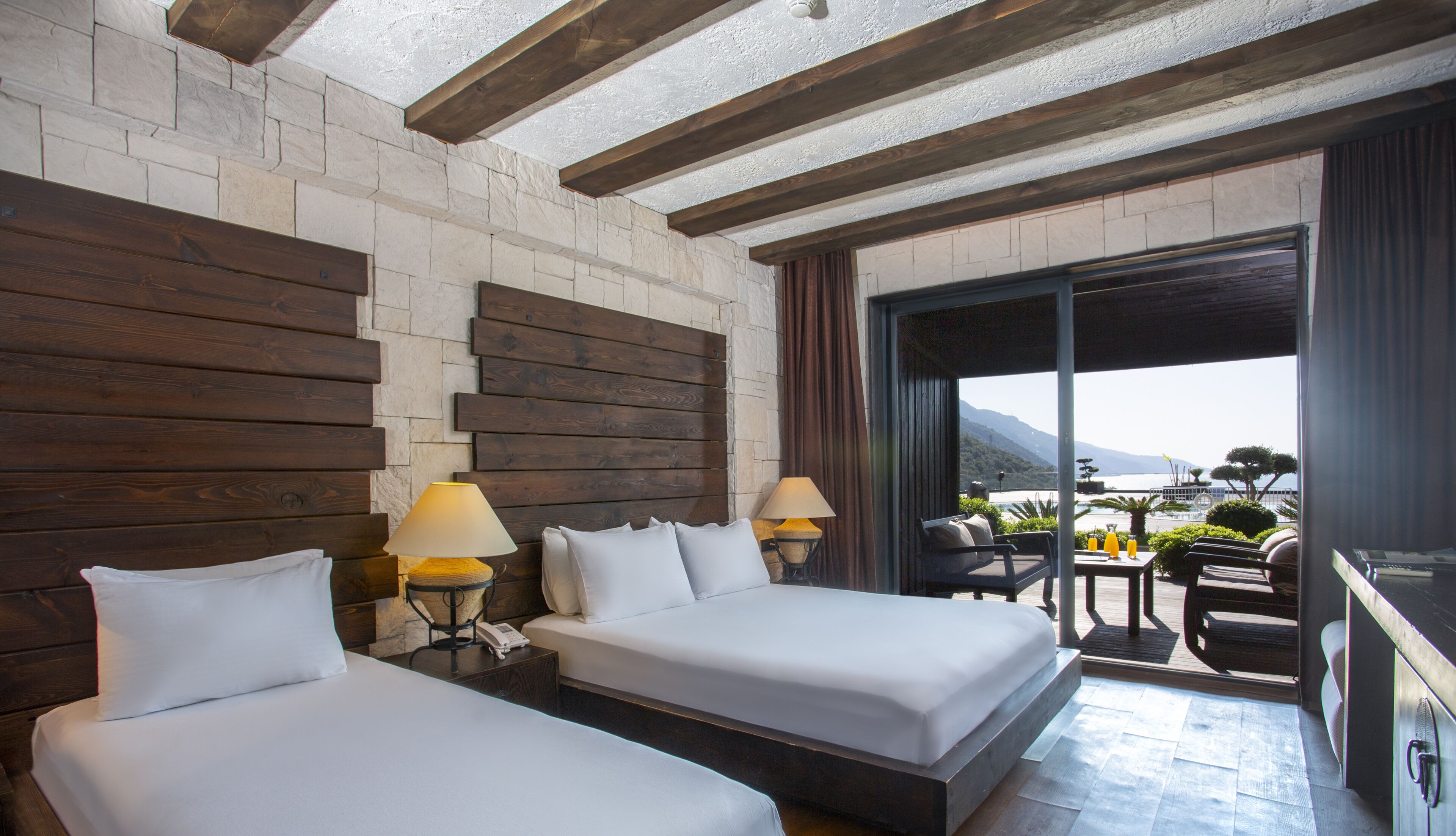 A hotel room with two beds, wooden headboards, a balcony with a table and chairs, and a view of the sea.