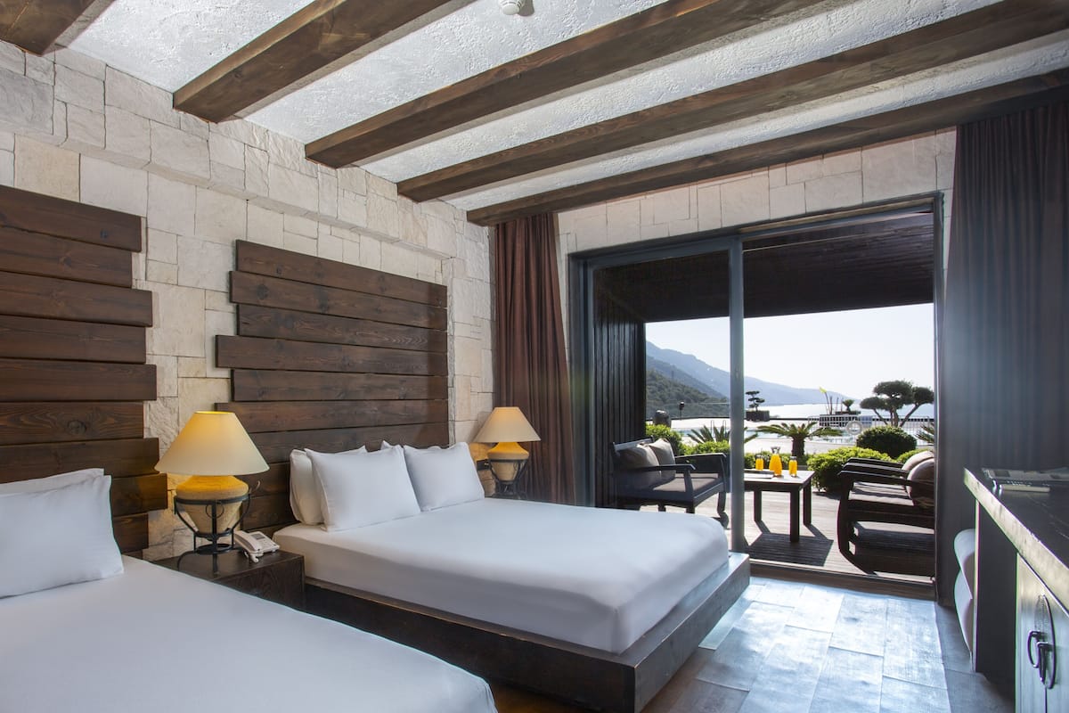 A hotel room with two beds, wooden headboards, a balcony with a table and chairs, and a view of the sea.