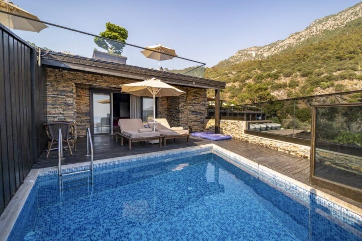 A rooftop pool with lounge chairs and umbrellas, surrounded by stone walls and a view of a mountainous landscape.