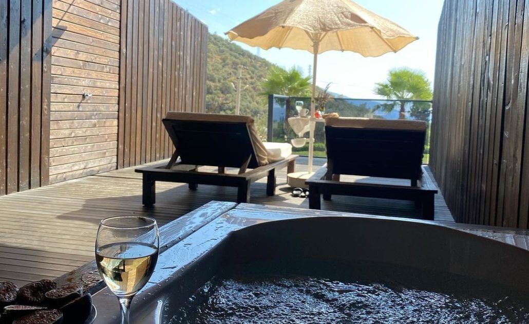Terraza de madera con dos sillones de descanso y una copa de vino, con vistas a una piscina.