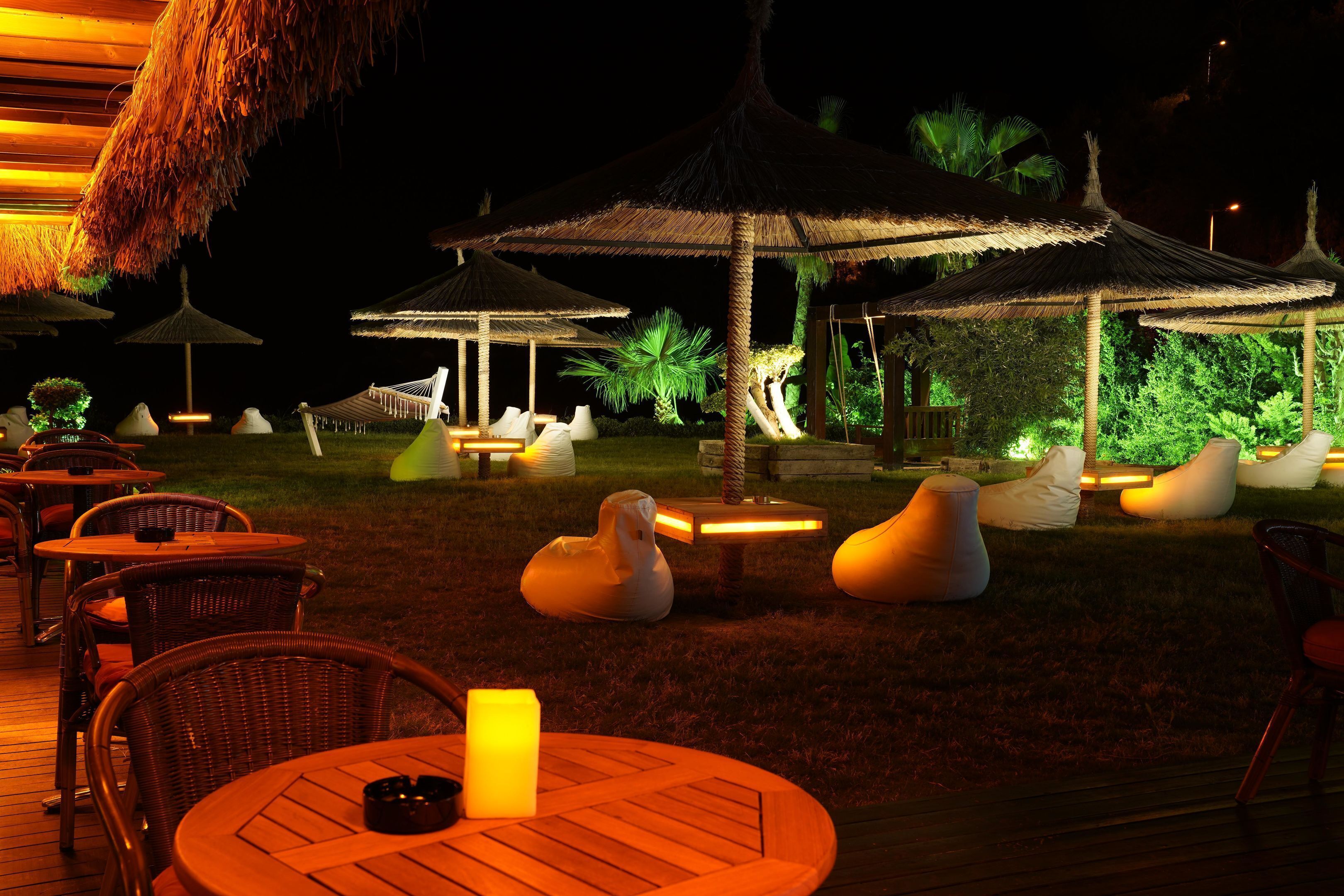 Un área de comedor al aire libre de noche con sombrillas de paja, mesas de madera y sillones de asiento tipo cojín blanco.