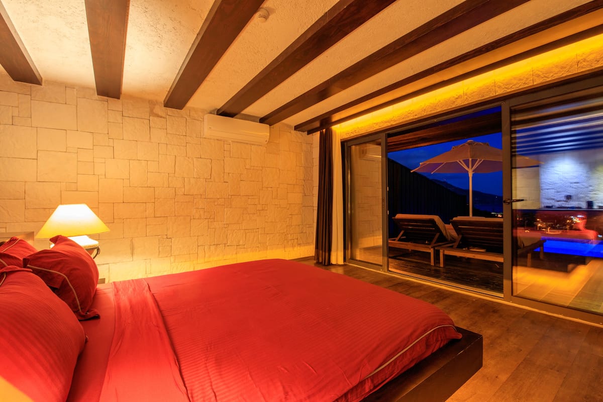 A bedroom with a large bed, a nightstand, a lamp, a sliding glass door leading to an outdoor seating area with a pool.