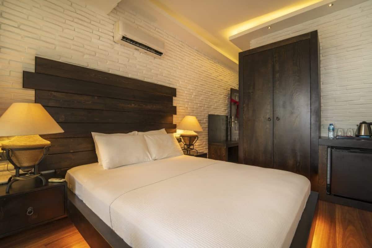 A hotel room with a large bed, two bedside tables with lamps, a wardrobe, and a small kitchenette area.