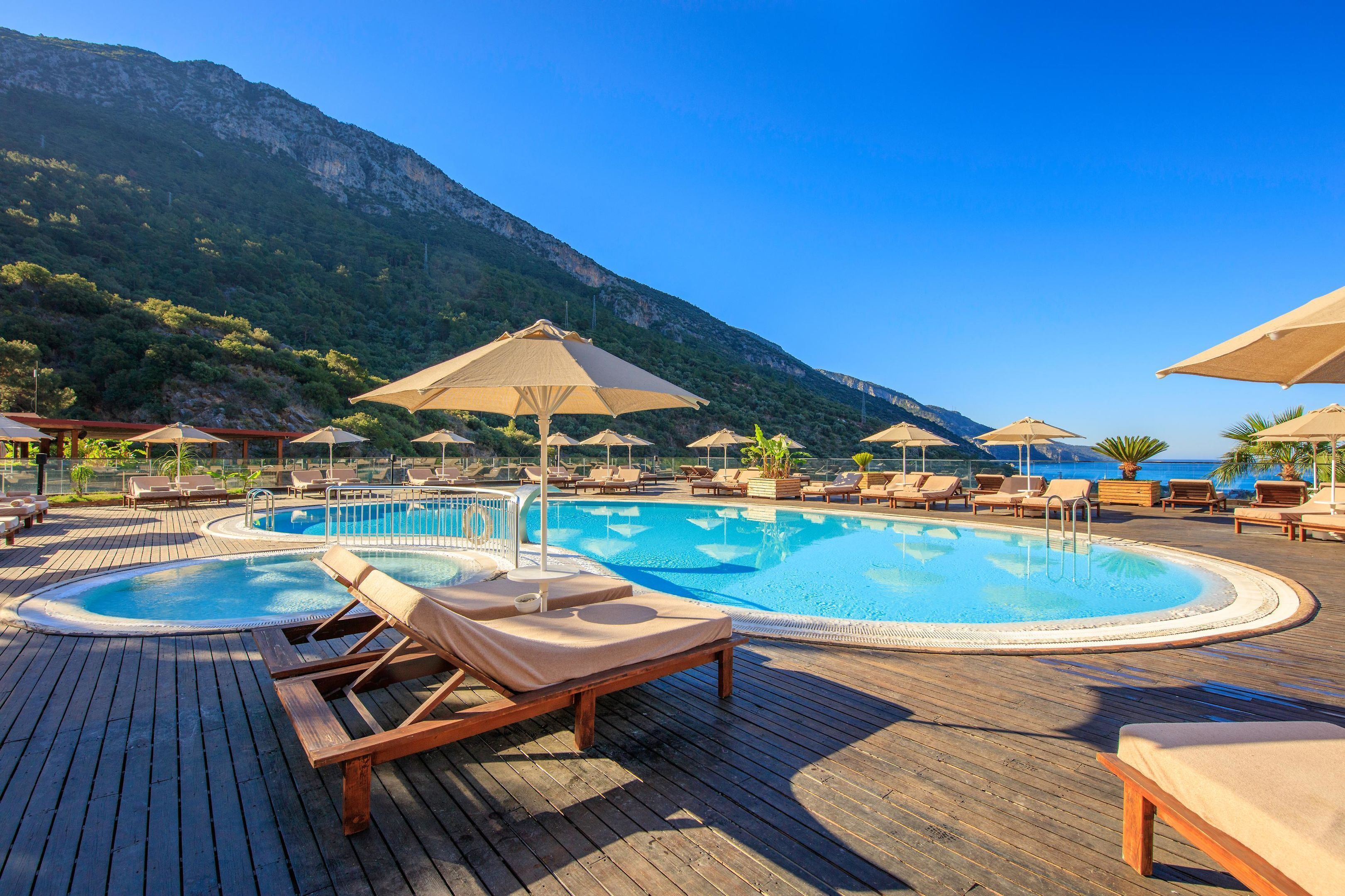 A pool area with lounge chairs and umbrellas, surrounded by a wooden deck and a mountainous backdrop.
