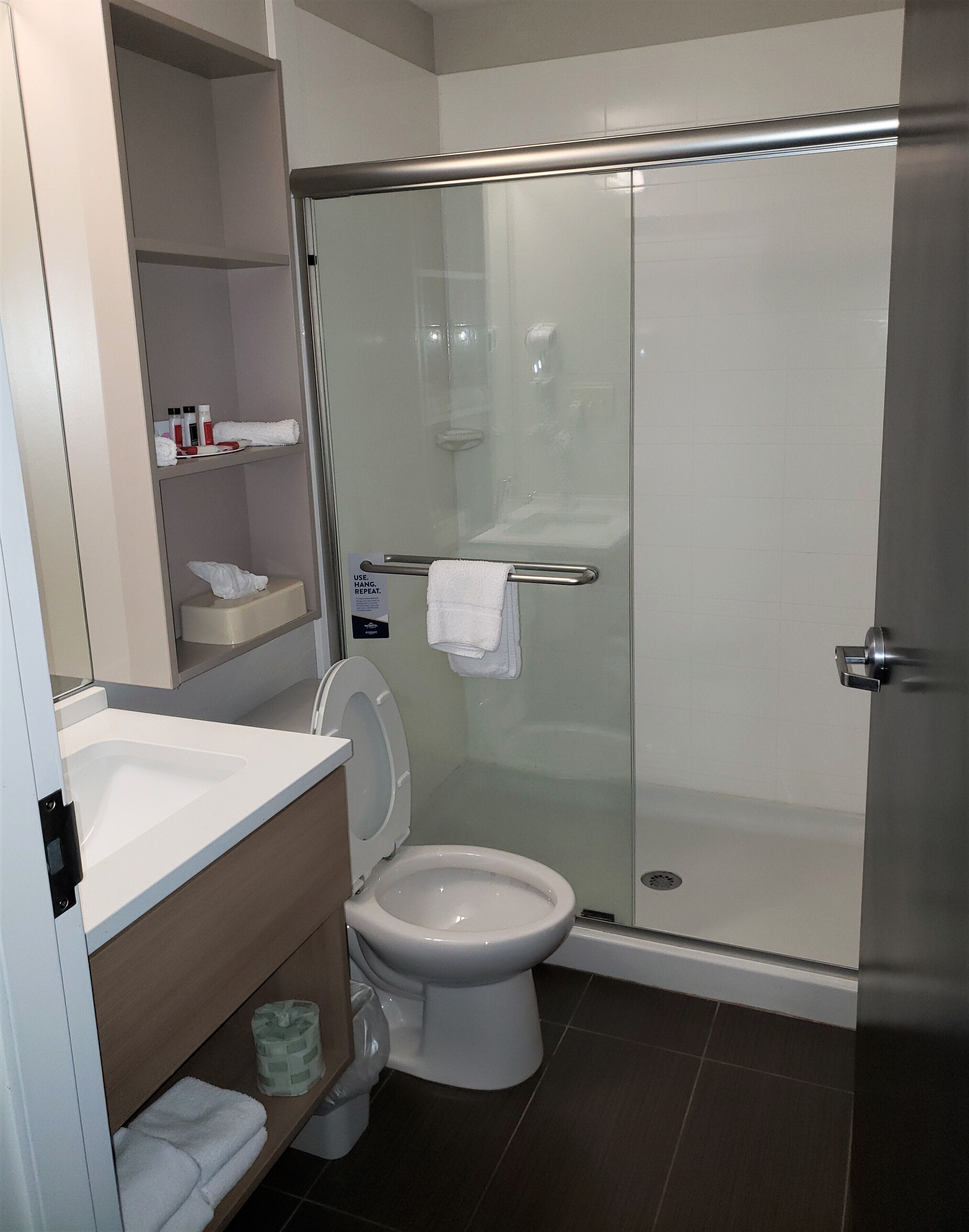 A modern bathroom with a glass-enclosed shower, a white toilet, a sink with a mirror, and a shelf with toiletries.