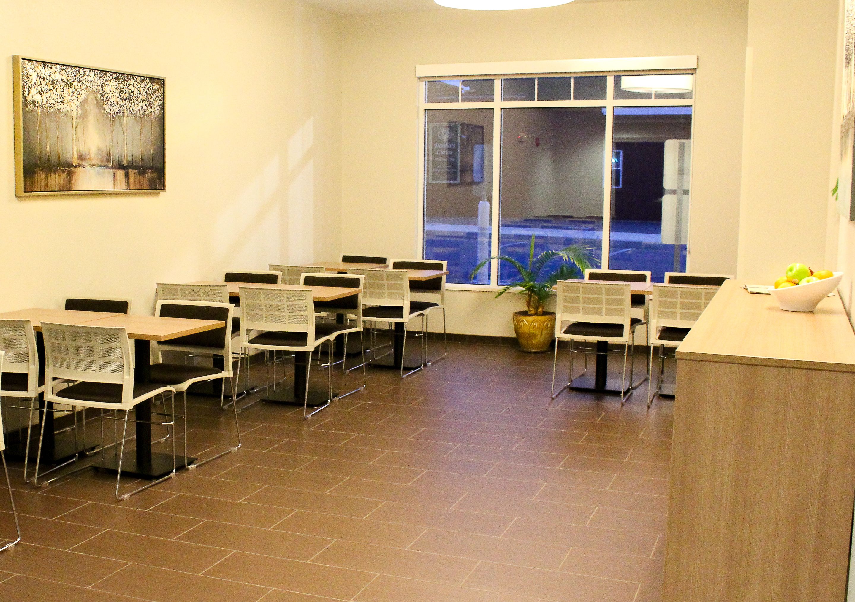 A conference room with white tables and chairs, a large window with a view of a parking lot, and a painting on the wall.