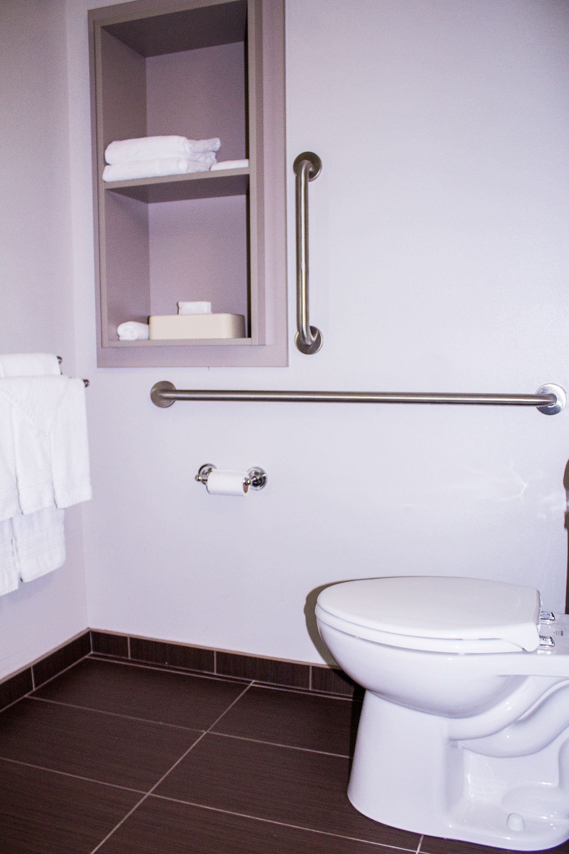 A bathroom with a toilet, a shelf with towels, a handrail, and a toilet paper holder.