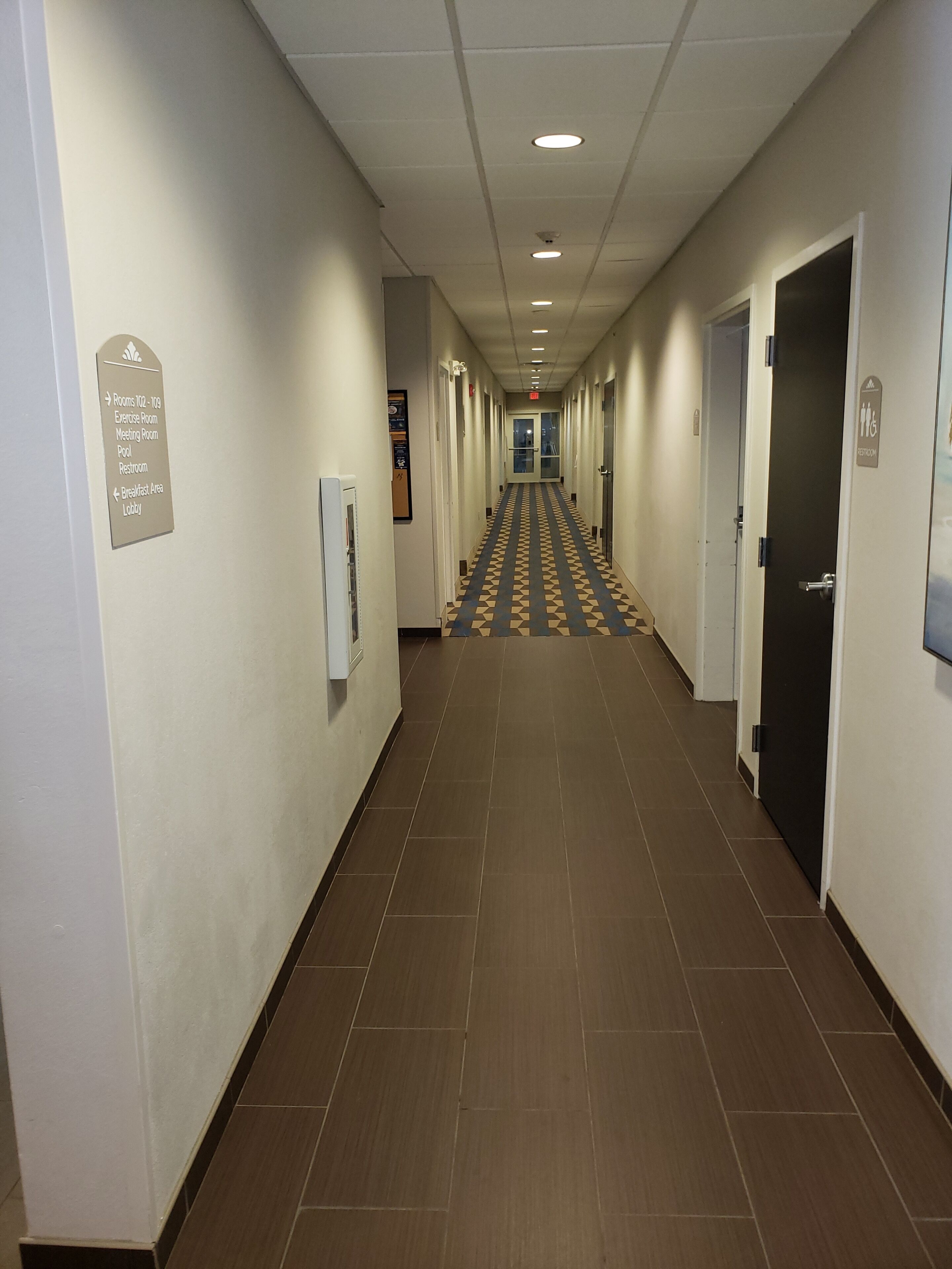 Un pasillo de hotel con un cartel que indica las habitaciones 102 a 106, incluyendo una sala de ejercicios, una sala de reuniones, piscina y baño.