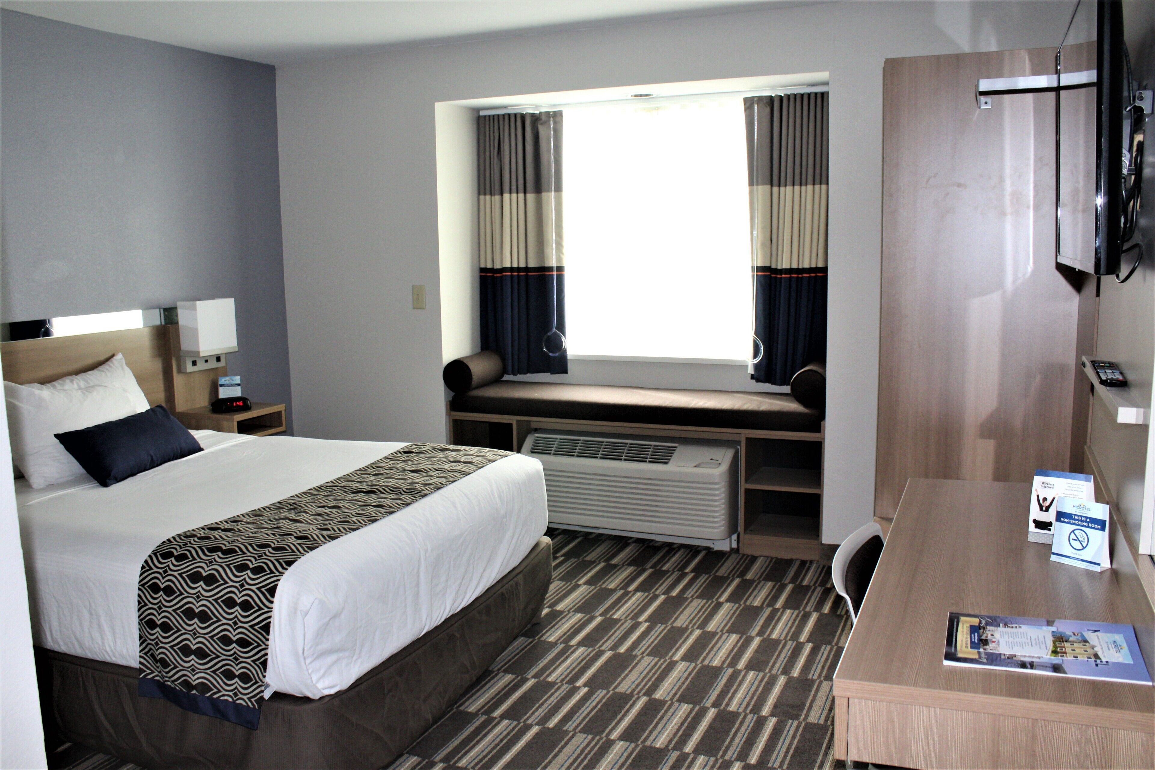 Habitación de hotel con una cama grande, un escritorio, una silla, una ventana con cortinas, un radiador y un televisor.