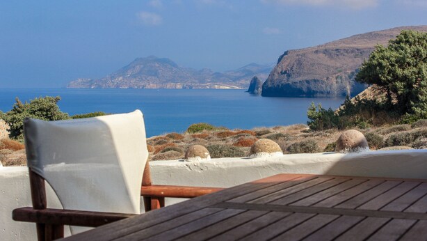 A wooden deck with a white cushioned chair and a table, overlooking a coastal landscape with cliffs and a body of water. - Avra Pahainas Hotel (Milos)