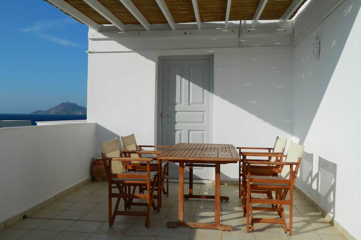 A covered outdoor patio with a wooden table and chairs, a white door, and a view of the sea and a mountain in the background.