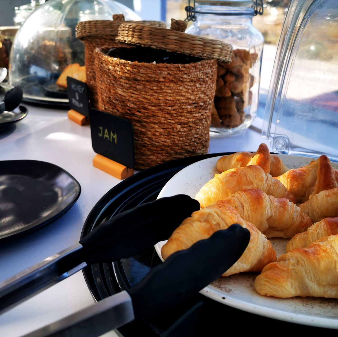 A display of croissants on a plate, a wicker basket labeled "JAM", and a glass jar with a label.