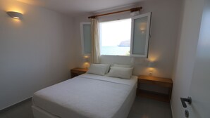 A bedroom with a bed, bedside lamps, a window with curtains, and a view of the sea. - Avra Pahainas Hotel (Milos)