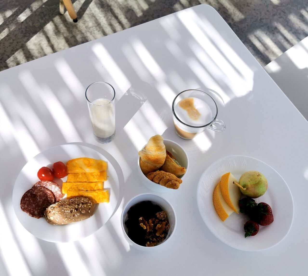 A white table with food items and drinks, including croissants, cheese, tomatoes, and a pear.