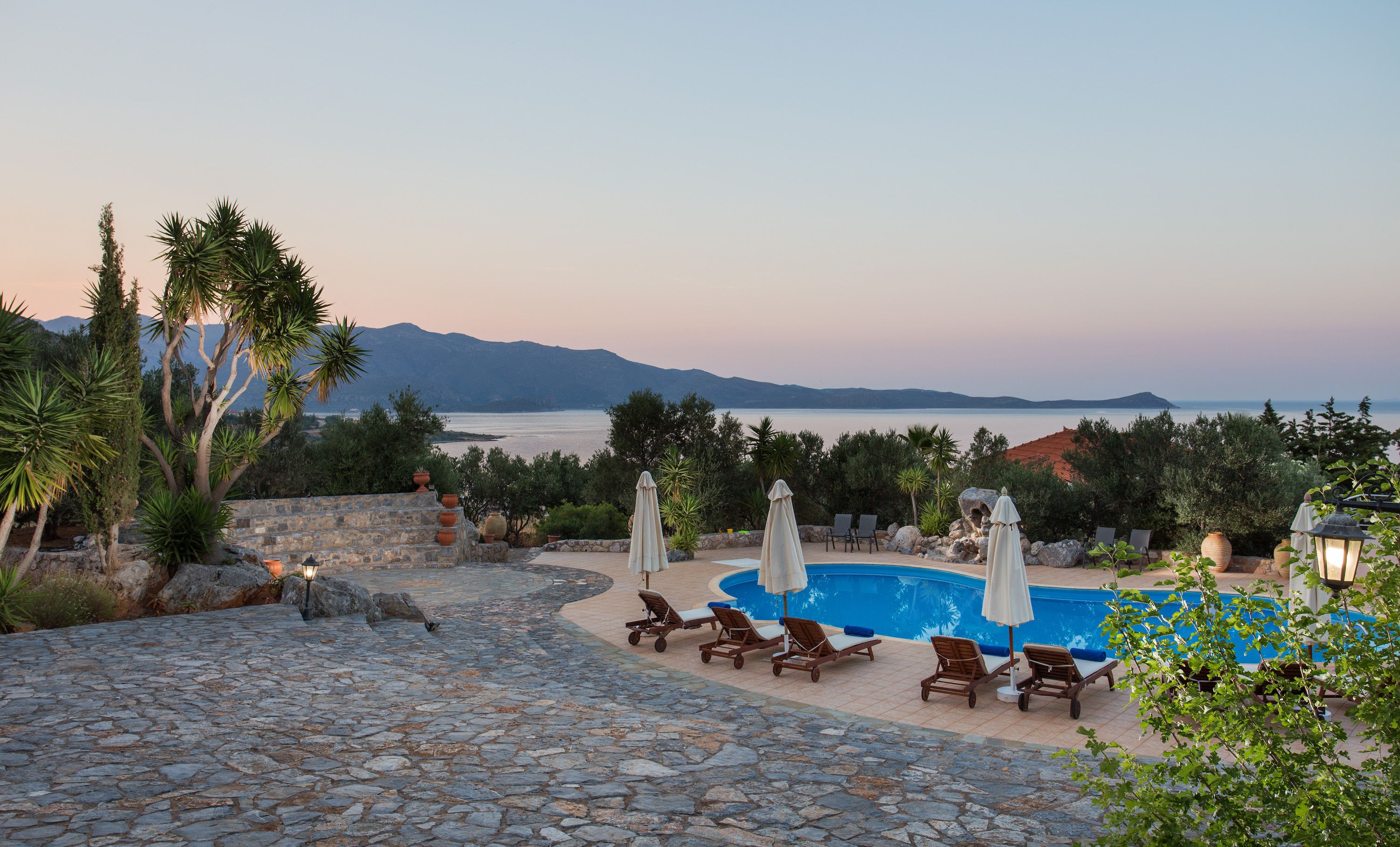 Un área de piscina con pavimento de piedra, sillones de descanso y sombrillas, con vistas al mar tranquilo y montañas a lo lejos.