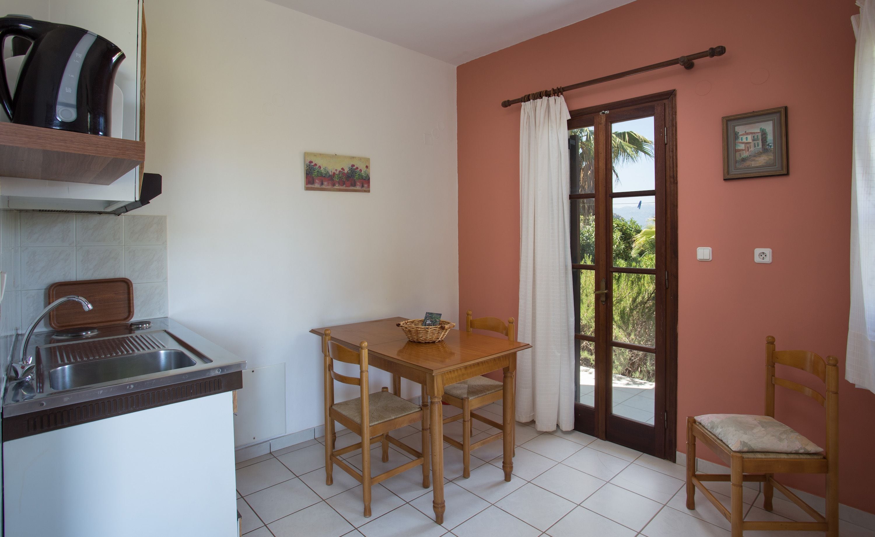 Une cuisine compacte avec un évier, une table en bois et des chaises. Une porte donne accès à un balcon d’où l’on peut admirer la verdure.