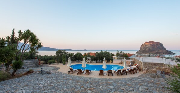 A pool area with lounge chairs and umbrellas, surrounded by stone steps and palm trees, overlooking a body of water and a mountain. - Monemvasia Village (Monemvasia)