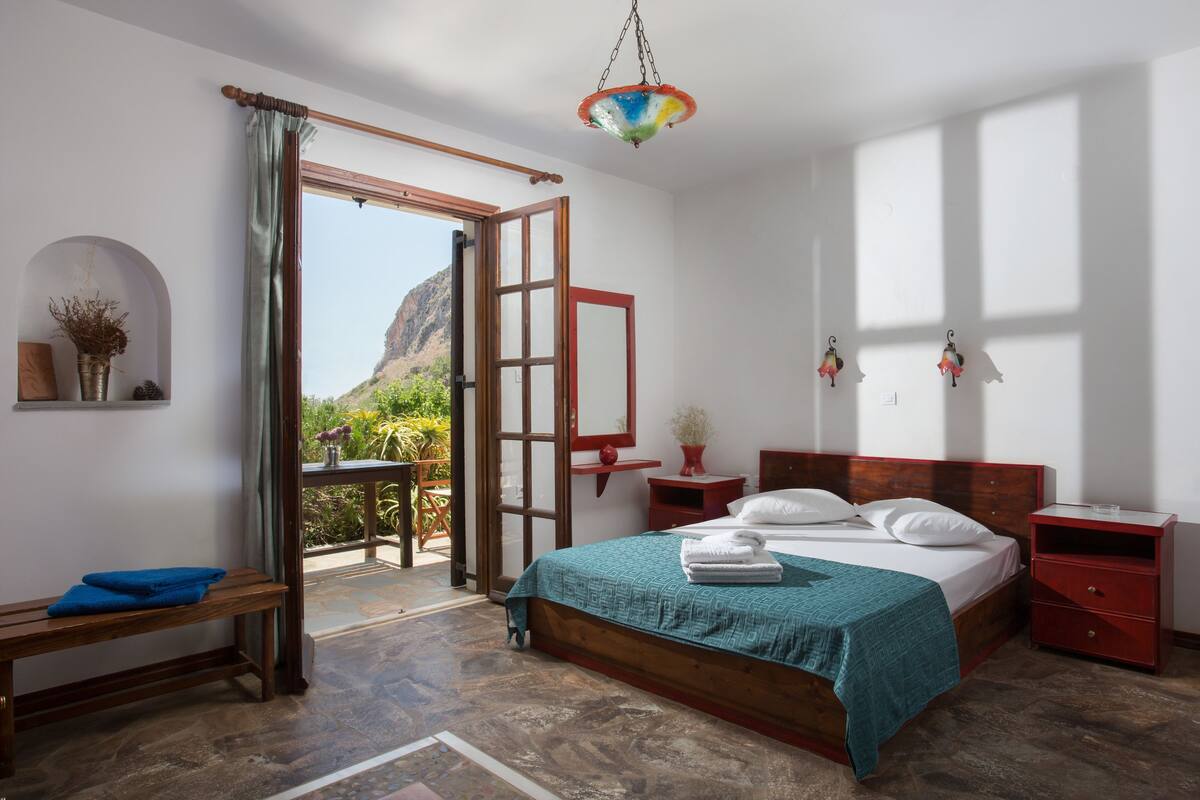 Ein Schlafzimmer mit Bett, einer Bank, einem Nachttisch, einer Nachttischlampe, einer Pendelleuchte und Blick ins Freie.