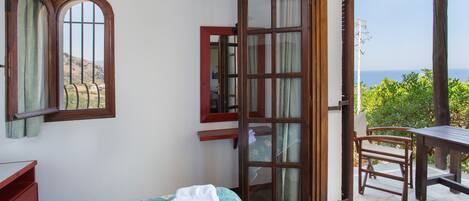 Un dormitorio con cama, escritorio, silla y vista al mar por una puerta abierta.