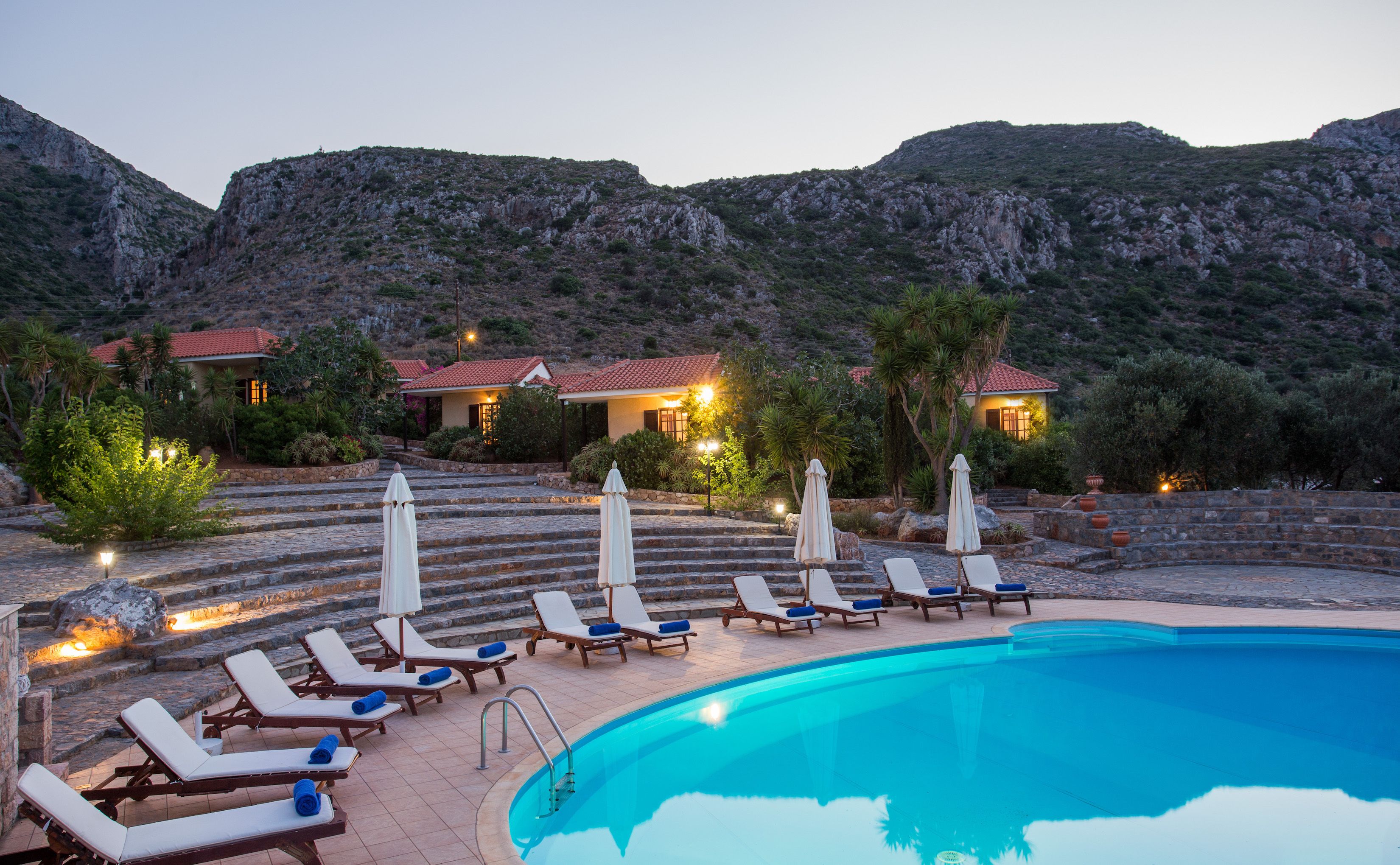 A poolside area with lounge chairs and umbrellas, a swimming pool, and a building with red-tiled roofs.