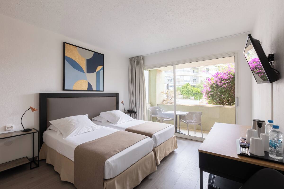 Habitación de hotel con dos camas, un escritorio, un televisor y un balcón con vistas a edificios y flores.