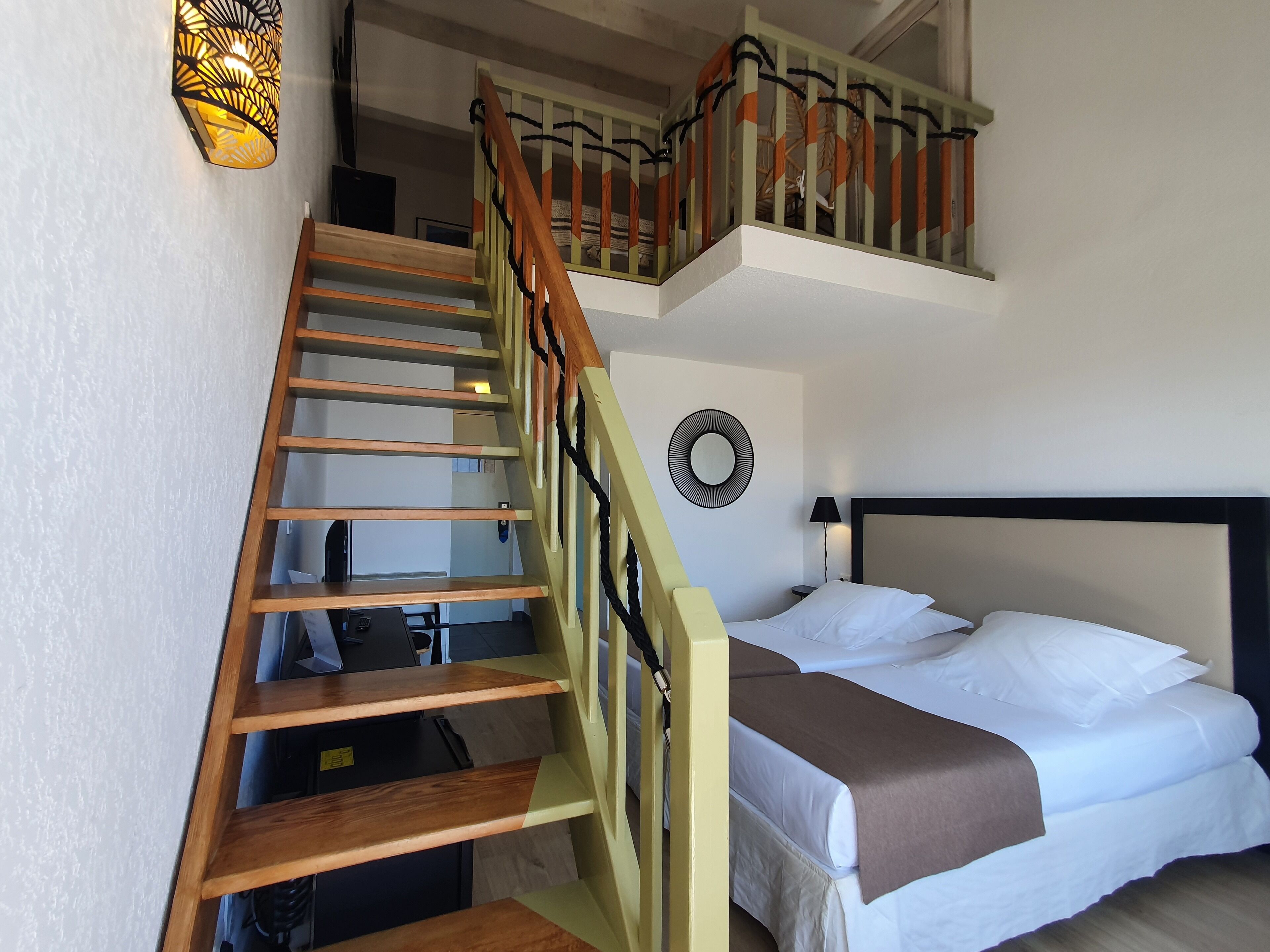 A bedroom with a bed, a bedside table, a lamp, a wall-mounted mirror, and a staircase with wooden steps and a railing.