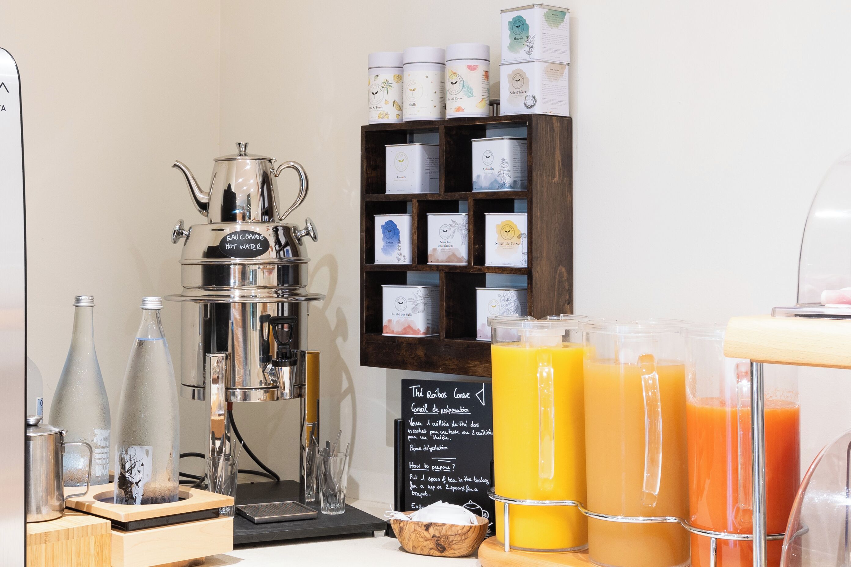 Um espaço com estação de café, incluindo máquina de café, prateleira com recipientes de chá e café, e um aviso com opções de bebidas.