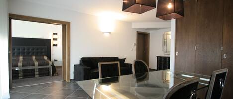 Uma sala de estar moderna com uma mesa de jantar de vidro, sofás escuros e uma cama grande com cabeceira.