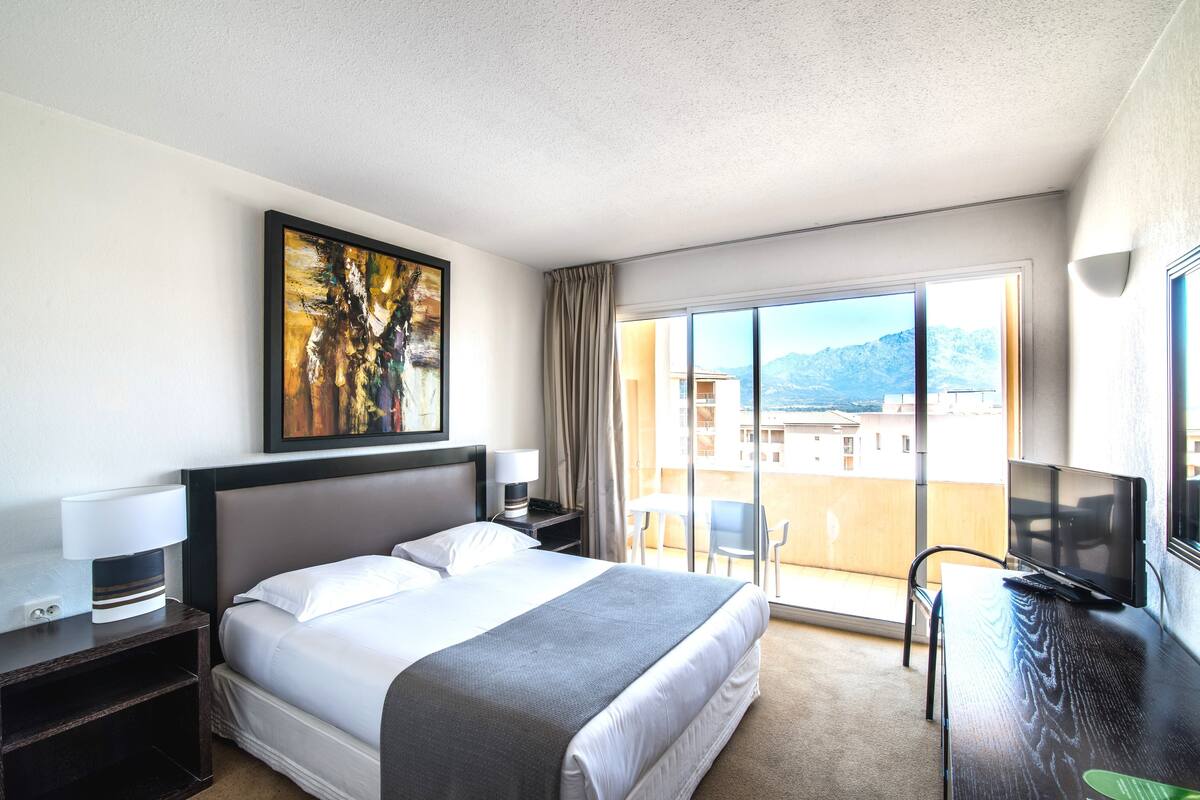 Habitación de hotel con cama, escritorio con televisor, pintura en la pared y balcón con vistas a edificios y montañas.