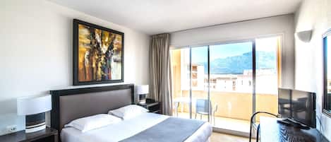 Habitación de hotel con cama, escritorio con televisor, pintura en la pared y balcón con vistas a edificios y montañas.