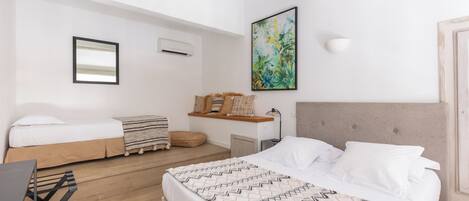 A modern bedroom with a large bed, a smaller bed, a desk, and a chair. There is a painting on the wall and a shelf with decorative pillows.