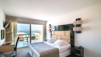 Habitación de hotel con cama, escritorio, silla, televisor y vista a las montañas y la playa.