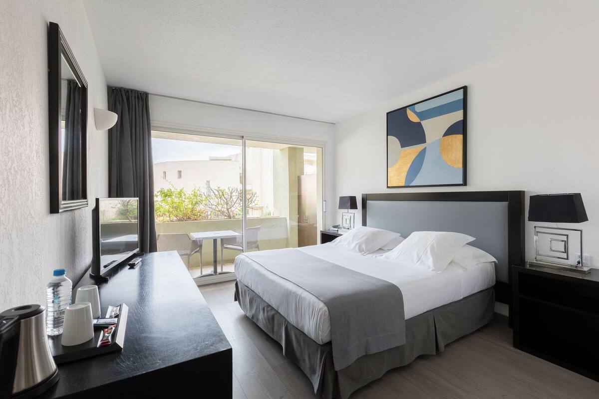 Habitación de hotel con cama, mesitas de noche, un escritorio con silla, un ventanal grande y un balcón.