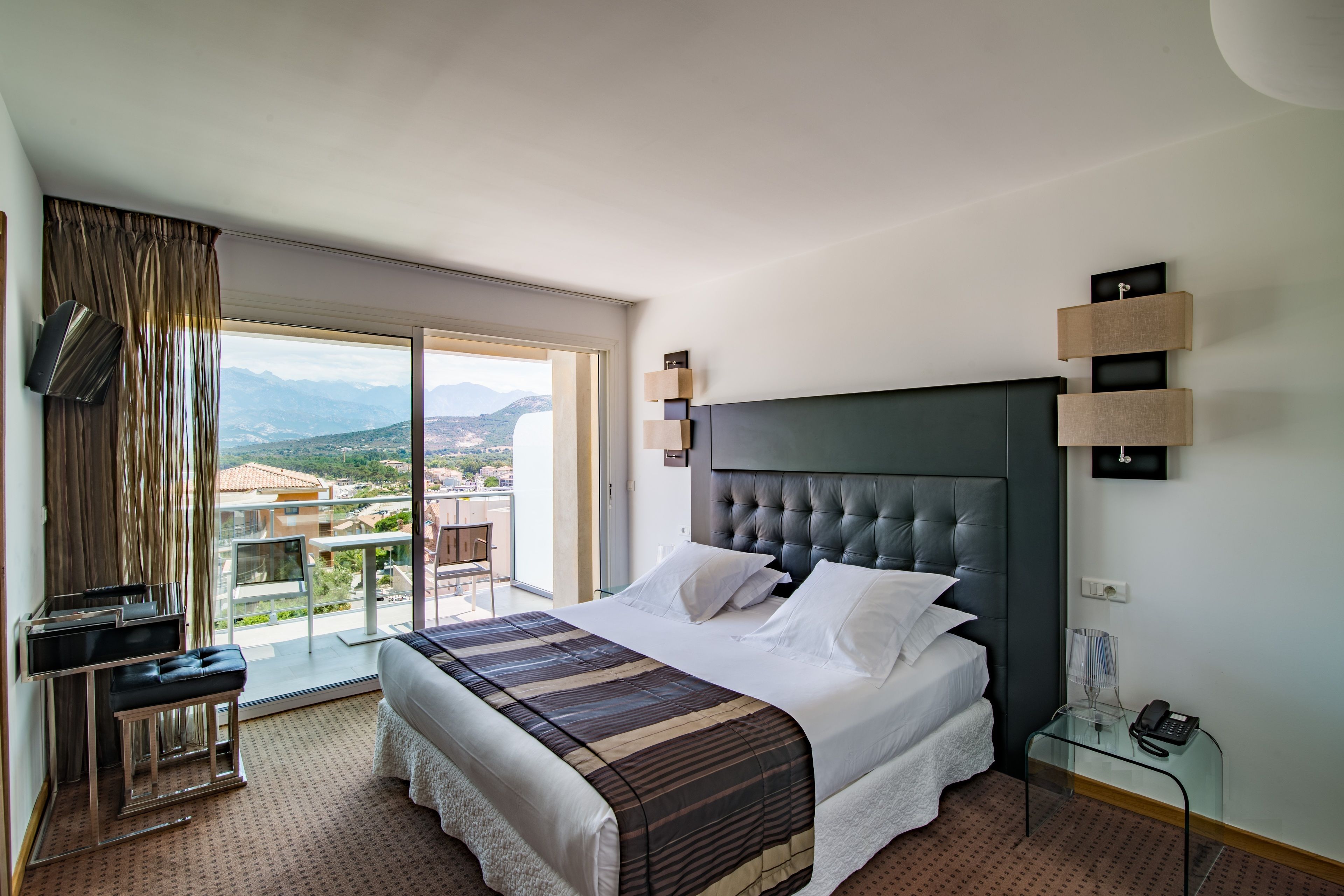 A hotel room with a large bed, a desk, a chair, a TV, and a balcony with a city view.