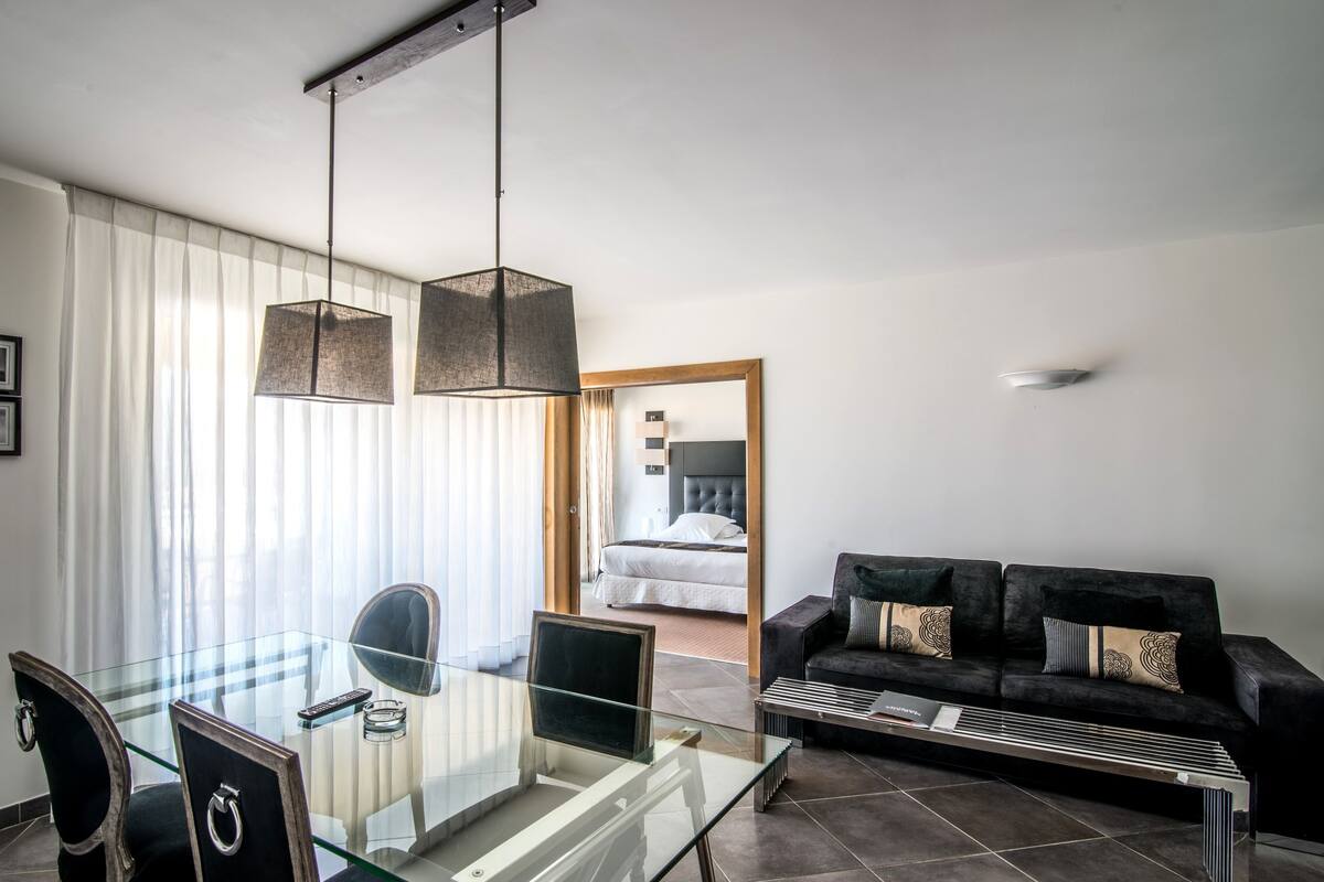 Una habitación de hotel moderna con una mesa de comedor de vidrio, un sofá negro y una cama con cabecera.