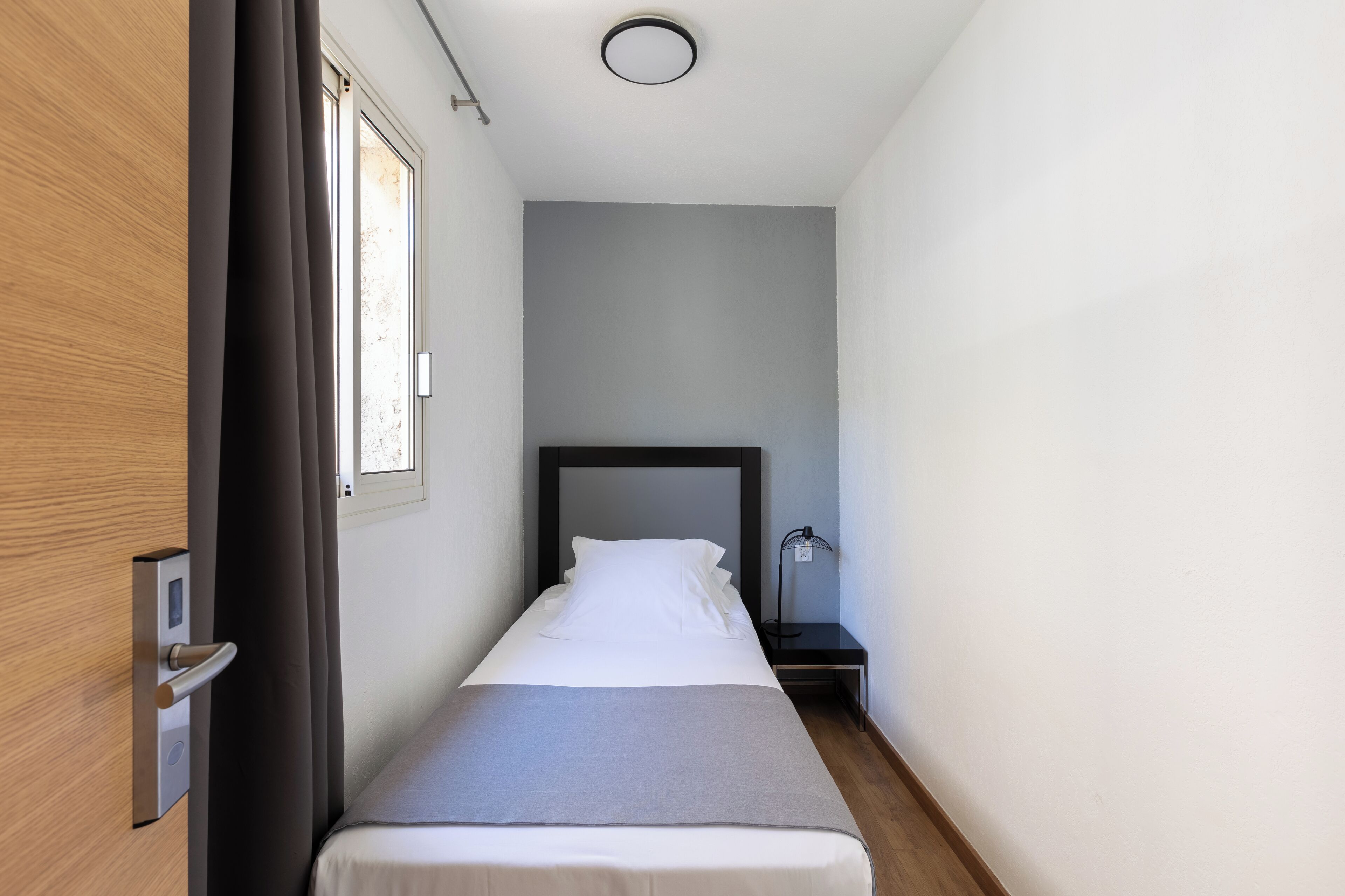 Une chambre d’hôtel étroite, avec un lit simple, une table de chevet, une lampe et une fenêtre.