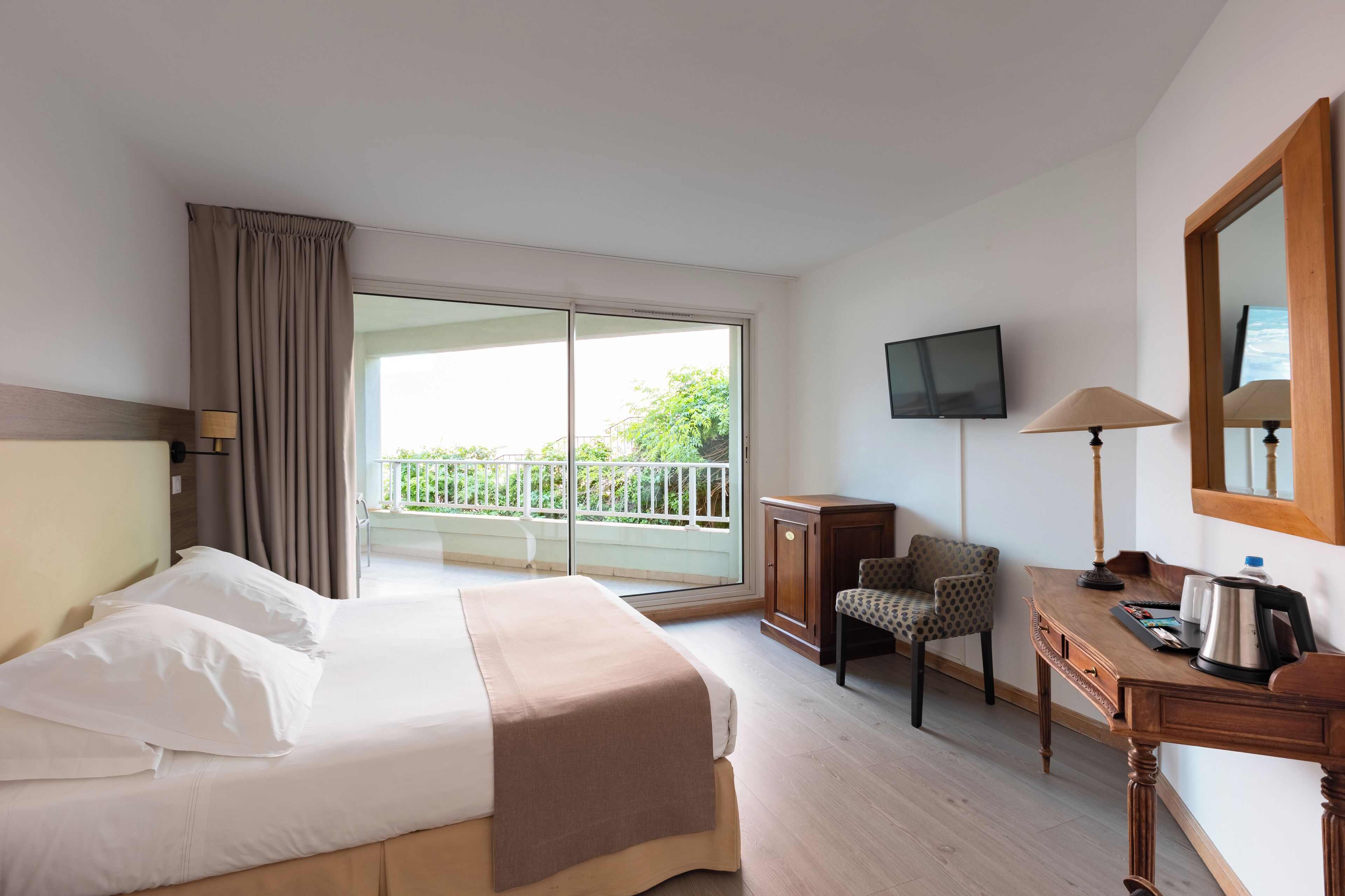 Une chambre d’hôtel comprenant un lit, un bureau avec une chaise, une télévision et un balcon donnant sur un espace verdoyant.