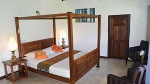 Uma cama de dossel com estrutura de madeira, uma mesa de cabeceira com luminária, uma cadeira e uma porta em um quarto com piso ladrilhado.