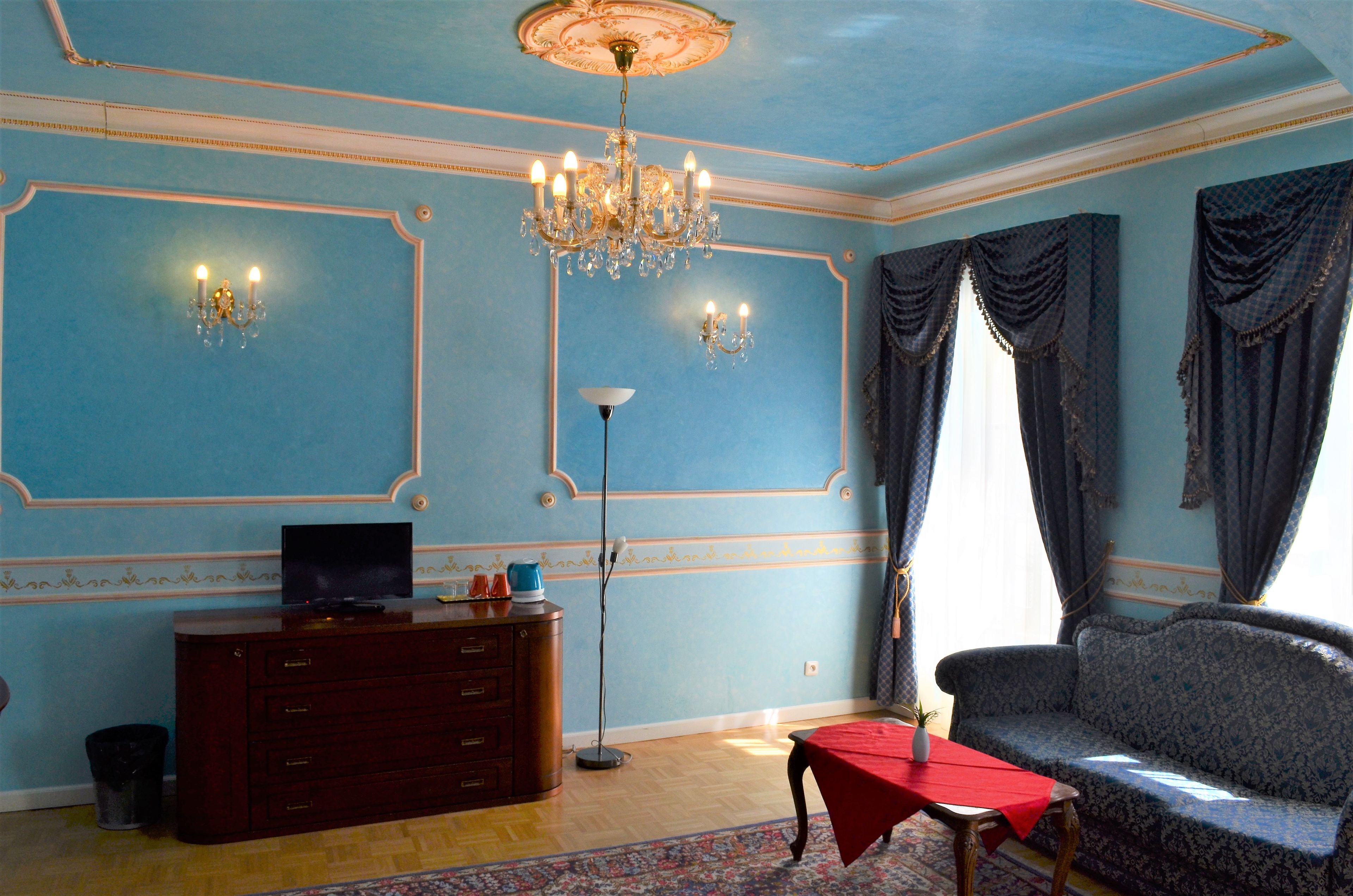 A room with a chandelier, a sofa, a television on a wooden cabinet, and a red table.